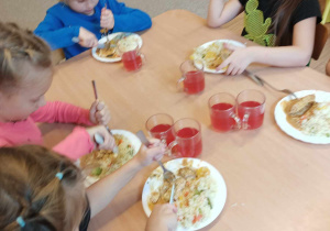 Dzieci siedzą przy stole i jedzą nożem i widelcem drugie danie.