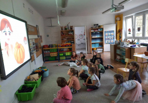 Dzieci siedzą na dywanie i oglądają prezentację multimedialną.