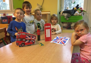Dzieci stoją wśród symboli Wielkiej Brytanii - czerwona budka telefoniczna, Flaga, czerwony piętrowy autobus, królewscy gwardziści