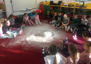 Dzieci obserwują, jak parujący suchy lód rozkłada sie po podłodze przypominając wyglądem chmury widziane z kosmosu