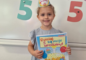 Marysia stoi na dywanie i pozuje do zdjęcia, trzymając urodzinowy dyplom