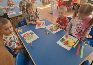 Grupa dzieci siedzi przy stoliku i wydziera papierem kolorowym kontur jabłka