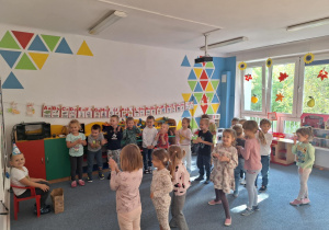 Dzieci stoją w półkolu na dywanie i śpiewają Wojtkowi sto lat