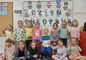 Chłopiec siedzi na krzesełku, trzymając w ręku napis -super chłopak, wkoło stoją dziewczynki z grupy