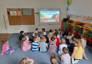 Dzieci siedzą na dywanie i oglądają film edukacyjny "Marta i dzień chłopaka"