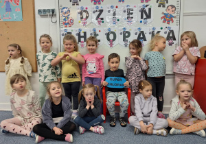 Chłopiec siedzi na krzesełku, trzymając w ręku napis -super chłopak, wkoło stoją dziewczynki z grupy