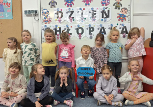 Chłopiec siedzi na krzesełku, trzymając w ręku napis -super chłopak, wkoło stoją dziewczynki z grupy