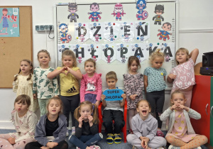 Chłopiec siedzi na krzesełku, trzymając w ręku napis -super chłopak, wkoło stoją dziewczynki z grupy