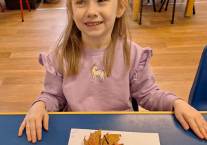 Dziewczynka siedzi przy stole i pokazuje swoją prace plastyczną