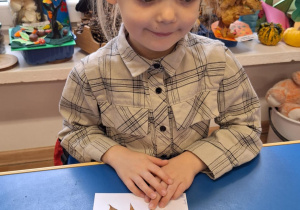 Dziewczynka siedzi przy stole i pokazuje swoją prace plastyczną