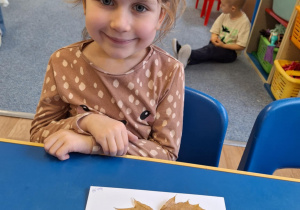 Dziewczynka siedzi przy stole i pokazuje swoją prace plastyczną