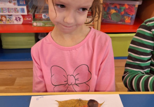 Dziewczynka siedzi przy stole i pokazuje swoją prace plastyczną