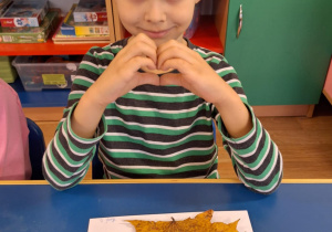 Chłopiec siedzi przy stole i pokazuje swoją prace plastyczną