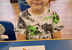 Chłopiec siedzi przy stole i pokazuje swoją prace plastyczną