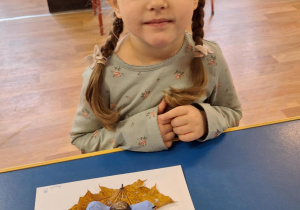 Dziewczynka siedzi przy stole i pokazuje swoją prace plastyczną