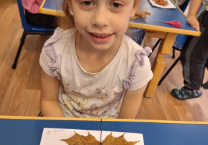 Dziewczynka siedzi przy stole i pokazuje swoją prace plastyczną