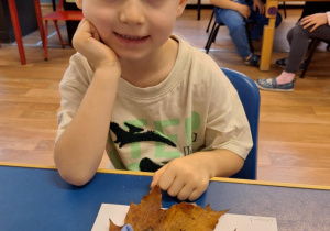 Chłopiec siedzi przy stole i pokazuje swoją prace plastyczną