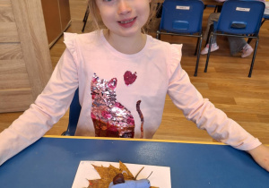 Dziewczynka siedzi przy stole i pokazuje swoją prace plastyczną