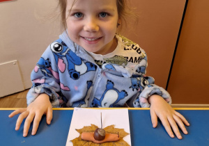 Dziewczynka siedzi przy stole i pokazuje swoją prace plastyczną