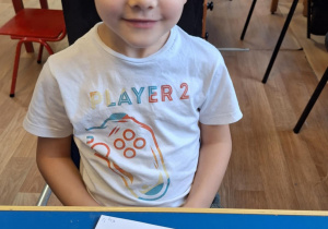 Chłopiec siedzi przy stole i pokazuje swoją prace plastyczną