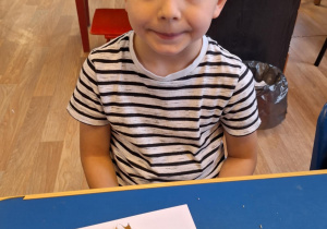 Chłopiec siedzi przy stole i pokazuje swoją prace plastyczną