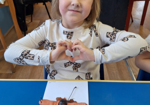 Dziewczynka siedzi przy stole i pokazuje swoją prace plastyczną