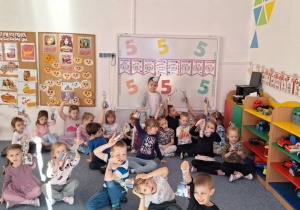 Dzieci siedzą na dywanie i pokazują trzymając w ręku upominek od Wojtka