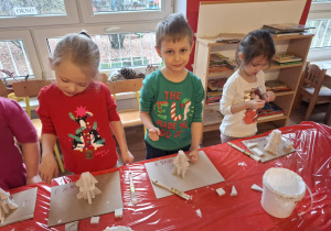 Dzieci stoją przy stole i sklejają gipsowe elementy choinki