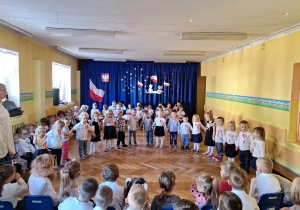 Dzieci ustawione w półkolu w strojach galowych z kotylionami pozują do zdjęcia