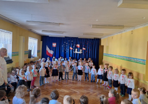 Dzieci ustawione w półkolu w strojach galowych z kotylionami pozują do zdjęcia