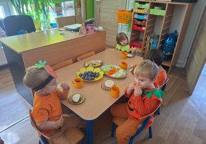 Dzieci przebrane za owoce i warzywa siedzą przy stole i jedzą zdrowe owoce i warzywa