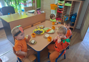 Dzieci przebrane za owoce i warzywa siedzą przy stole i jedzą zdrowe owoce i warzywa