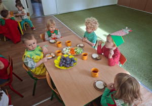 Dzieci przebrane za owoce i warzywa siedzą przy stole i jedzą zdrowe owoce i warzywa