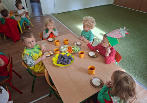 Dzieci przebrane za owoce i warzywa siedzą przy stole i jedzą zdrowe owoce i warzywa