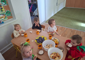 Dzieci przebrane za owoce i warzywa siedzą przy stole i jedzą zdrowe owoce i warzywa