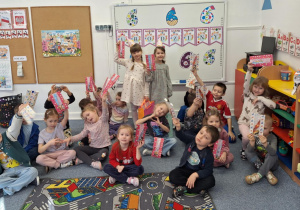 Cała grupa dzieci siedzi na dywanie i pokazuje upominek od Justyny i Marceliny