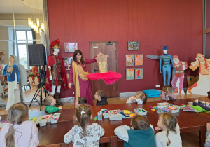 Grupa dzieci siedzi przy stole i słucha pani prowadzącej pokazującej stroje