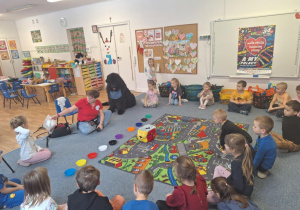 Grupa dzieci siedzi na dywanie i słucha pani prowadzącej zajęcia