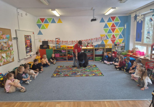 Dzieci siedzą na dywanie i słuchają pani prowadzącej zajęcia