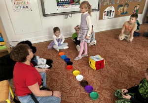 Dziewczynka stoi przed kolorowymi podstawkami, panią i psem