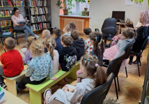 Dzieci siedzą na krzesłach i słuchają bajki czytanej przez panią bibliotekarkę