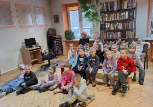 Dzieci siedzą na krzesłach i słuchają bajki czytanej przez panią bibliotekarkę