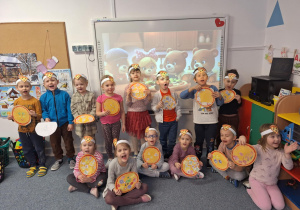 Grupa dzieci stoi na dywanie i pozuje do zdjęcia ze zrobioną przez siebie prace plastyczną-pizzą