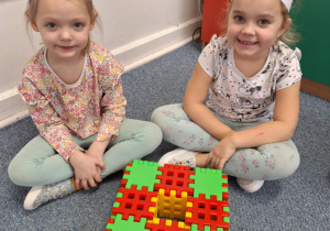 Dziewczynki siedzą na dywanie i pokazują pączka zrobionego z klocków