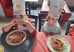 Dziewczynki siedzą przy stole i pokazują pizzę przez siebie zrobioną
