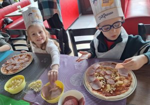 Dwoje dzieci siedzi przy stole i robi swoje pizze