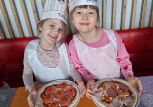 Dziewczynki siedzą przy stole i pokazują pizzę przez siebie zrobioną