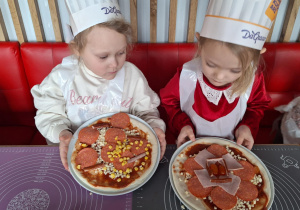 Dziewczynki siedzą przy stole i pokazują pizzę przez siebie zrobioną