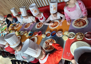 Grupa dzieci siedzi przy stole i robią swoją pizzę