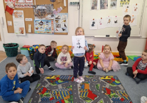 Dziewczynka stoi na dywanie i pokazuje dzieciom obrazek z kotem z bajki